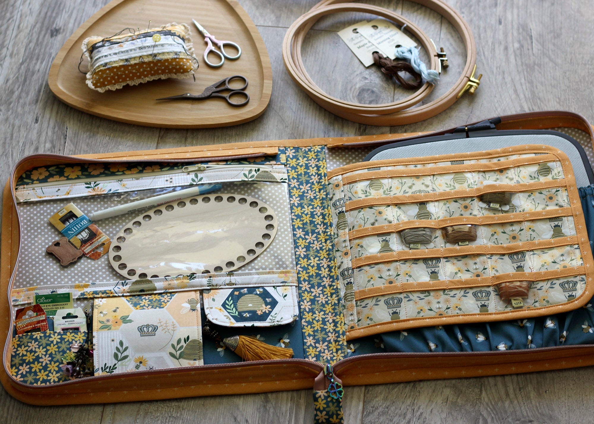 A wooden embroidery storage case opened flat on a gray wood floor, filled with fabric hoops, thread, scissors, and sewing tools. Two matching wooden embroidery hoops and a small fabric pin cushion sit above the case.